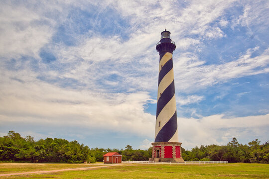 A daytime landscape of the Cape Hatteras Lighthouse on the beach under a cloudy blue sky on the Outer Banks in North Carolina.
