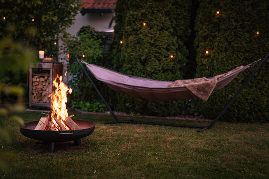 Hammock with fairy lights in garden. Cozy evening atmosphere with a campfire in a fire bowl. Background for leisure activities for outdoors.