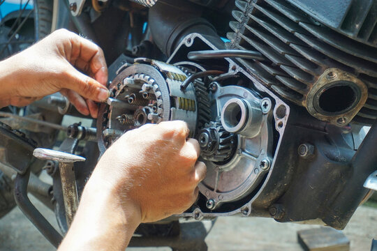The process of manually repairing or servicing a motorcycle engine by a mechanic. The mechanic's hands are seen removing or installing a cover component near the cylinder block of a two-stroke engine.