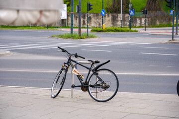 Naklejka premium Black mountain bike secured with a chain lock to a metal post on a city sidewalk, with an empty crosswalk and road infrastructure in the background, symbolizing urban transport and waiting