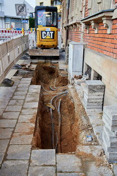 A yellow CAT mini excavator next to a deep trench with exposed utility cables on a sidewalk in Germany.