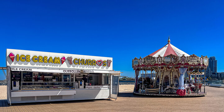 Ostend, Belgium - April 30, 2026: Ice cream and churros kiosk next to a traditional vintage carousel on the seaside promenade in Ostend, Belgium, on a sunny spring day with clear blue sky.