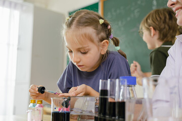 A young girl carefully conducts a science experiment with liquids in a classroom, focusing intently while a teacher and another student observe in the background. © Minttita