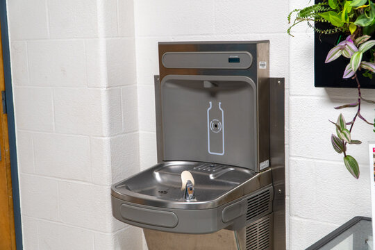 Modern stainless steel water fountain and bottle filling station with a 'Bottles Saved' counter against a white brick wall, promoting eco-friendly hydration and plastic waste reduction.