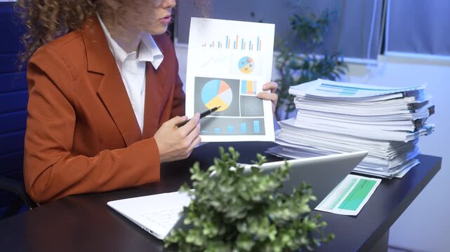 Businesswoman in office presenting analytics report with charts, laptop on desk, large stack of paperwork and green plant foreground; corporate finance, workload and strategy concept.