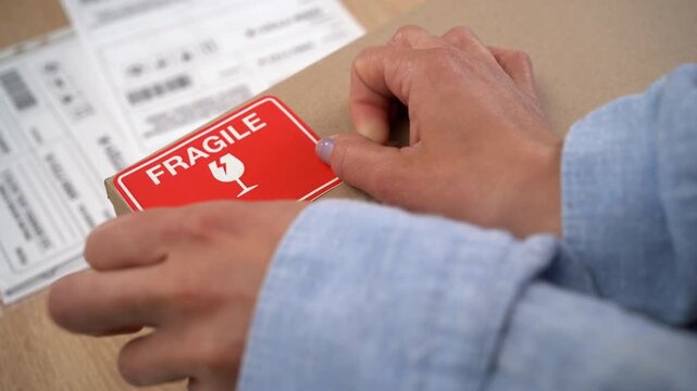 close-up of female hands put-on red shipping label to cardboard box. label the word FRAGILE