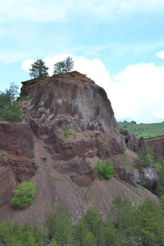 A dramatic landscape of the former volcano Racoș in Romania, featuring red rocky cliffs, green forest, and a bright blue sky. Unique volcanic formations create a stunning natural scene.
