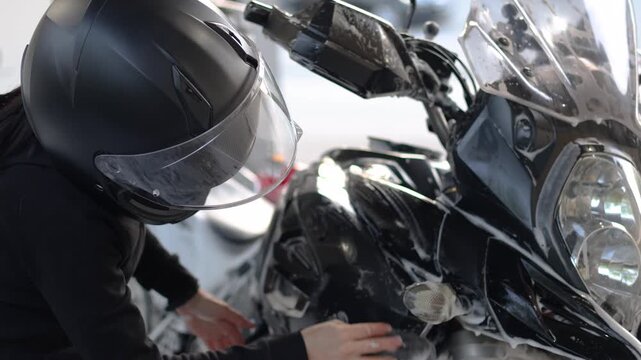 A woman in a black jumpsuit and helmet washes her motorcycle with a rag after applying foam at a self-service car wash. Slow motion footage of a woman takes care of her motorcycle.