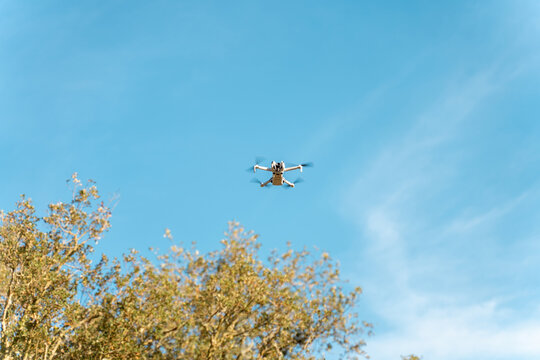 Small white drone flying in clear blue sky over trees
