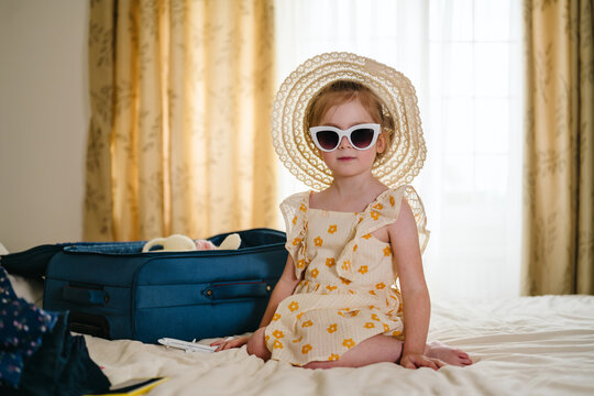Girl in hat and sunglasses sitting on bed with suitcase for travel