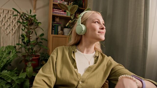 Handheld zoom in shot of relaxed young blonde Caucasian girl holding smartphone and enjoying listening to music in headphones while resting on sofa in green living room with macrame wall decor