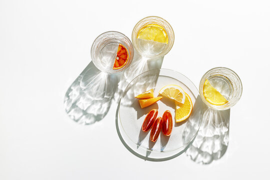 Clear Glasses and plate as summer still life with shadow and sunlight reflection on white table, close up glass cups of infused fruit water, slises of lemon, orange, tangerine, healthy summer drink
