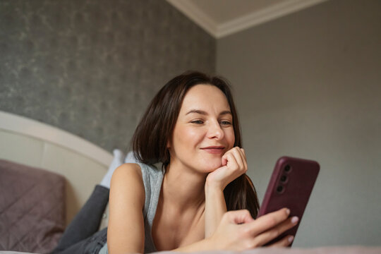 Smiling woman using a smartphone while relaxing on a bed at home