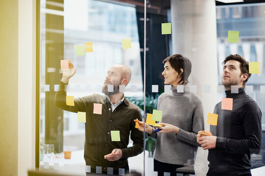 Businessman sticking adhesive note on glass wall and discussing with coworkers in office