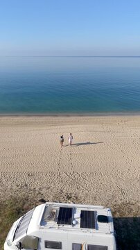 Two friends walk along the sandy shores of Chalkidiki, Greece, soaking in the sun and enjoying the peaceful atmosphere. A camper van parked nearby adds to the holiday vibe.