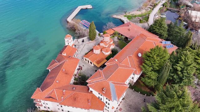 A cinematic aerial drone shot moving forward over the Monastery of Saint Naum with a smooth pan down to focus tightly on the red-tiled Byzantine domes of the Church of the Holy Archangels