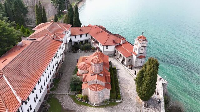 A cinematic aerial drone pull-back from the Church of Saint Naum, revealing the fortified monastic courtyard, the tall bell tower, and the vibrant turquoise waters of Lake Ohrid