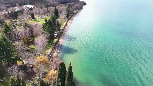 A wide cinematic aerial drone pull-back from the Church of Saint Naum, revealing the fortified monastic courtyard, the adjacent lakeside hotel complex, and the vast Lake Ohrid shoreline