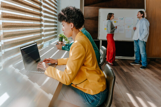 Programmer coding on laptop while team discusses in sunny office