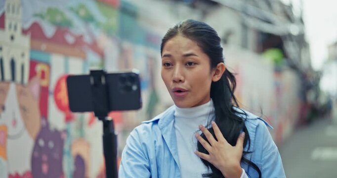 Young Asian woman records a video vlog using a smartphone mounted on a gimbal. She speaks engagingly to the camera while standing in an urban alley with colorful street art.