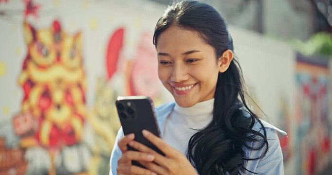 Cheerful young Asian woman with long dark hair smiles while looking at her smartphone. She stands outdoors against a colorful, artistic graffiti wall in a lively urban setting.