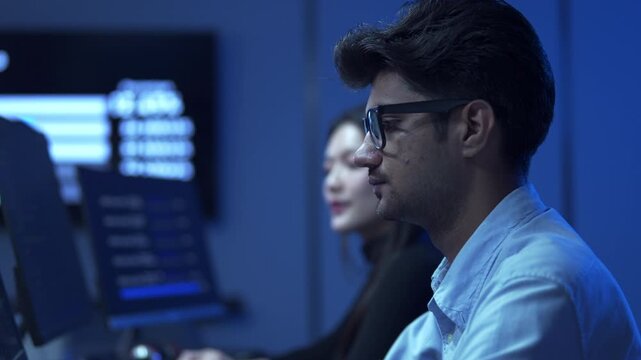 Young male software engineer writing program code on multiple desktop screens while his colleague working in the background in a modern office.
