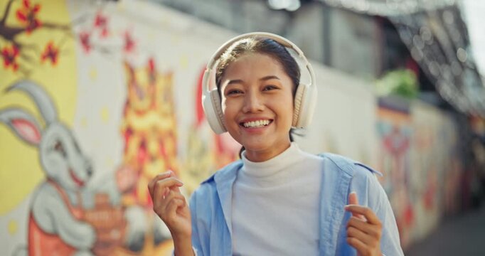 Carefree young Asian woman in a denim shirt closes her eyes and smiles while listening to music through white wireless headphones, dancing on a colorful sunlit urban street.