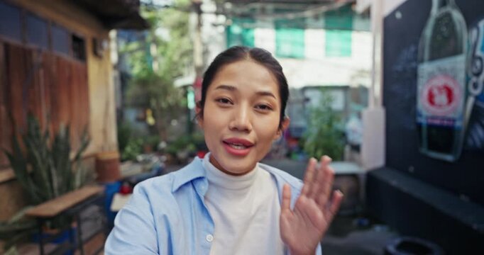 Pretty young Asian travel vlogger in a denim shirt looks directly into the camera and gestures while filming a friendly video log in a quiet old town alley with motorbikes.