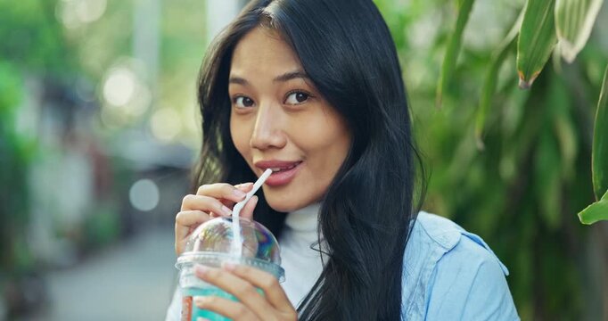 Beautiful young Asian woman with long black hair sips a colorful iced drink through a straw while standing near tropical plants at a cafe during a relaxing summer travel day.
