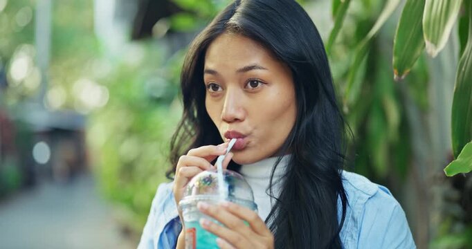 Elegant young Asian woman with long flowing black hair drinks a refreshing iced beverage through a straw while standing among tropical foliage on a sunny summer travel day.