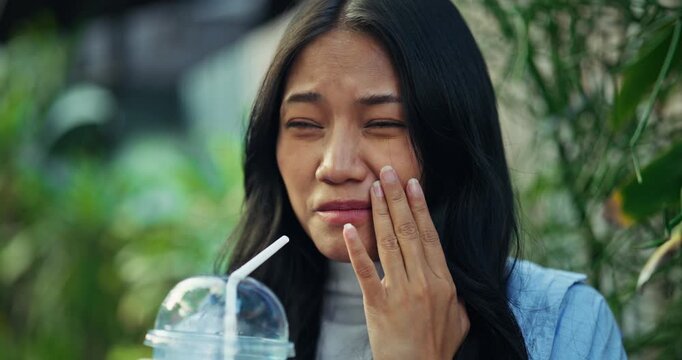 Funny candid moment as a young Asian woman holds her face and reacts with discomfort to brain freeze headache after sipping an icy cold beverage on a hot summer travel day.