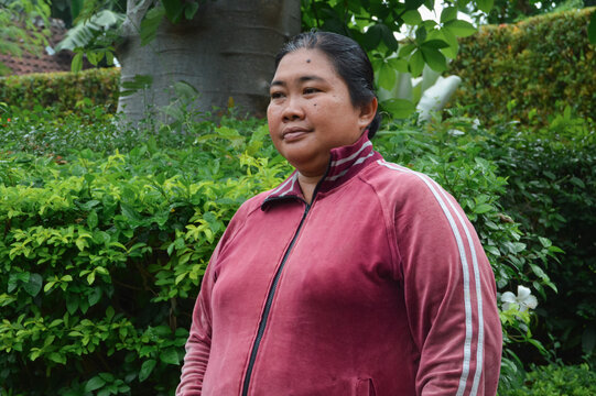 An Asian woman in a red track jacket stands thoughtfully with a background of lush green bushes and a large tree trunk.