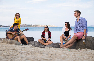 Fototapeta premium Friends enjoy a casual day at the beach, with one person playing an acoustic guitar while others sit and socialize, a serene coastal landscape in the background.