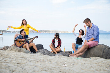 Obraz premium Friends enjoy a casual day at the beach, with one person playing an acoustic guitar while others sit and socialize, a serene coastal landscape in the background.