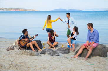 Fototapeta premium Friends enjoying a day at the beach, one playing guitar while others dance and socialize, capturing a carefree summer moment