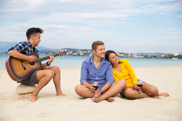 Obraz premium A young man plays an guitar while two friends sit and chat beside him on a sunny beach. The trio enjoys a casual afternoon by the ocean, with a scenic coastline and cloudy sky in the background.