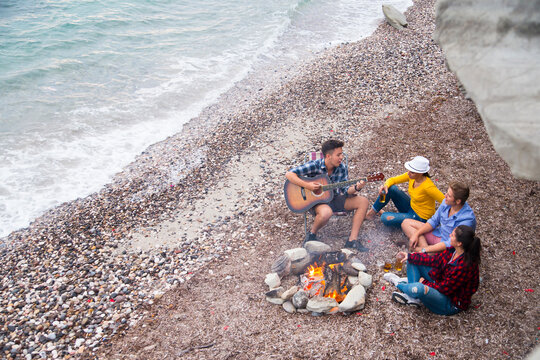 Group of friends sitting on beach with guitar playera campfire in a  outdoor setting, sitting on beach with guitar player.