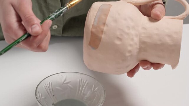 4K Video Slow Motion artist uses a wide studio paintbrush to apply thick glaze to a bisque ceramic bowl for an artistic finish. Artisan Glazing Ceramic Bowl with a Brush