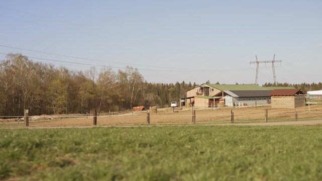 Calm countryside view featuring a lush green field in the foreground, with a farm, horse paddocks, and a dense forest in the background under a clear blue sky on a tranquil sunny spring day