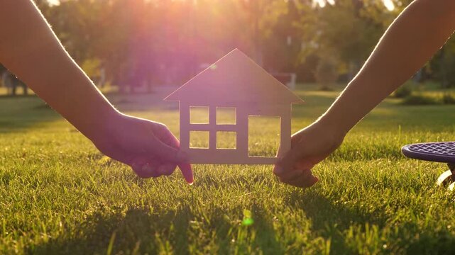 Hands hold miniature house model in sunlight. Outdoor family activity symbolizes home dreams and aspirations. Positive atmosphere showcases creativity and connection. Family unity in new home.