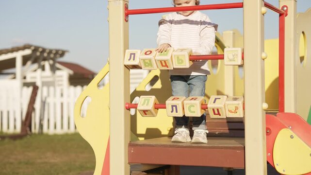 Cute toddler girl playing with large abacus style alphabet blocks on a colorful outdoor playground, spinning the cubes with cyrillic letters on a sunny day to develop fine motor skills