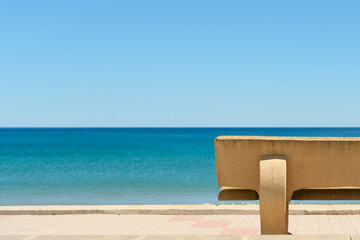 Fototapeta premium Modern concrete bench overlooking a calm Mediterranean beach and turquoise sea under clear blue sky. Minimal coastal composition with peaceful summer atmosphere and copy space.