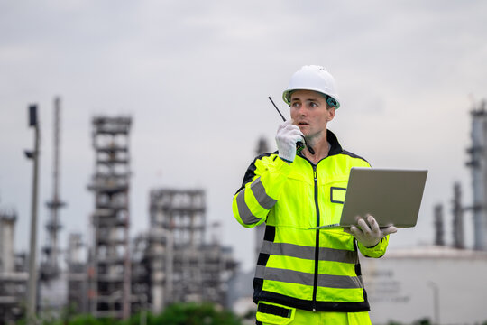 Professional male industrial engineer speaking on walkie talkie holding laptop reporting at petrochemical refinery plant for sustainable corporate oil and gas manufacturing business management.