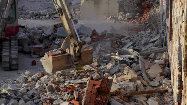 Excavator bucket lifts broken bricks, concrete and debris at a demolition site, showing heavy machinery clearing construction waste and dusty rubble.