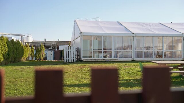 Large white wedding marquee situated in an industrial area on a green lawn with a clear blue sky, viewed from behind a wooden fence during a sunny day, showcasing an outdoor event venue. 4K