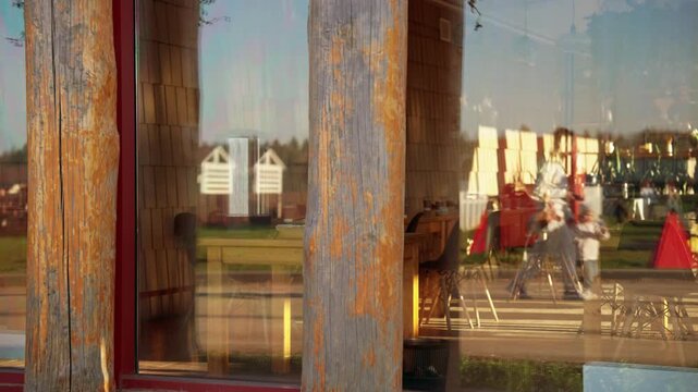 Slow motion reflection in a window of a mother and her young child walking together past rustic wooden beams, enjoying a sunny day at a vibrant outdoor country fair or market. Slow motion, 4K