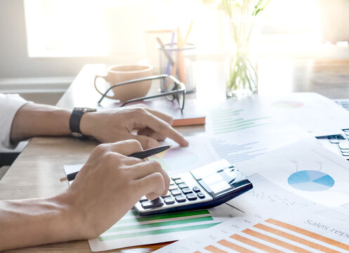 Person calculates finance using calculator and charts. Fingers press keys on calculator. Papers show colorful graphs and data. Natural light from window illuminates desk. Image ideal for business, acc
