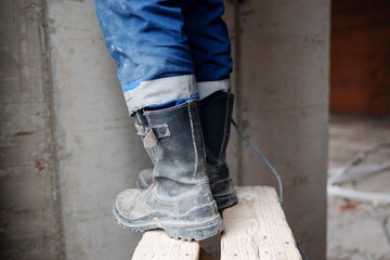 A construction worker in protective boots stands on a wooden step stool next to a concrete pillar with electrical cables on the floor in the background. © Aigul Sabirova