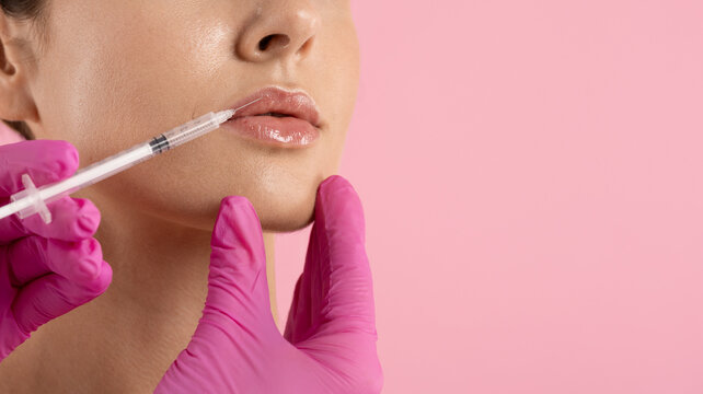 Closeup of young woman receiving hyaluronic acid lip filler injection by doctor hands in pink gloves, lip augmentation cosmetic procedure on pink background