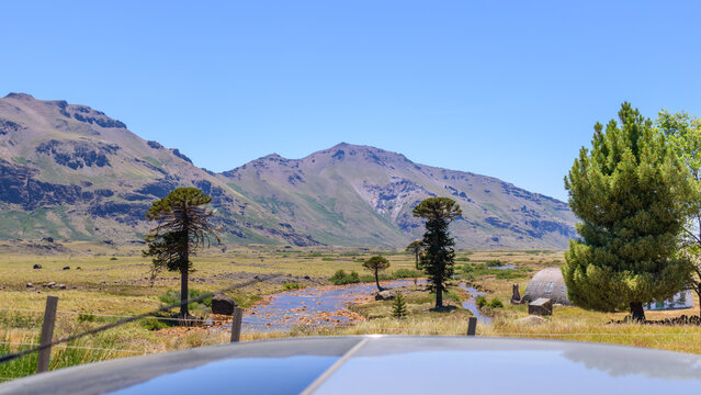 Paisaje rural en Caviahue con el r&iacute;o Agrio, &aacute;rboles de araucarias, un hangar met&aacute;lico y monta&ntilde;as bajo un cielo azul despejado, Neuqu&eacute;n, Patagonia, Argentina.